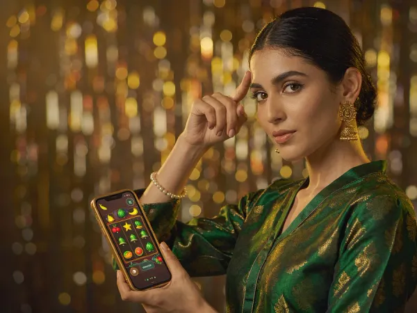 m444 Woman in emerald and gold holds a phone showing a fruit and bell slot game